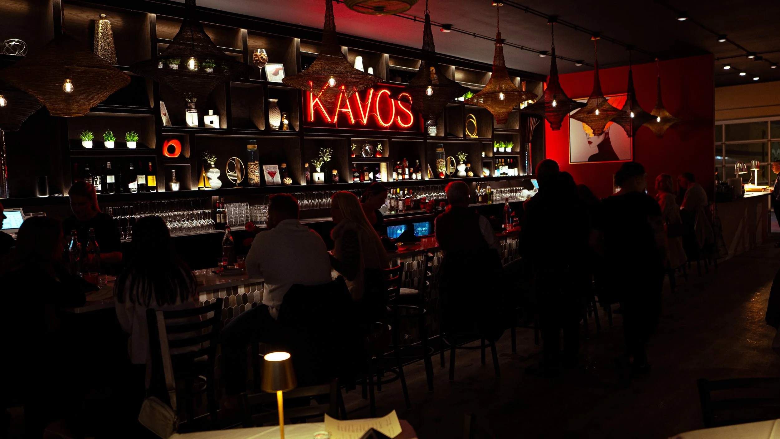 Indoor bar scene at night with people sitting and standing, shelves displaying bottles of alcohol, a neon sign that says 'KAVOS,' and decorative lighting fixtures hanging from the ceiling.