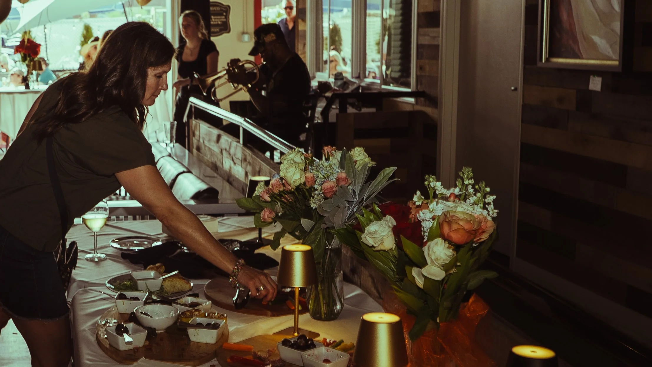 A woman preparing food at a table with a bouquet of roses and other flowers, set in a restaurant or cafe with large windows and a view outside.