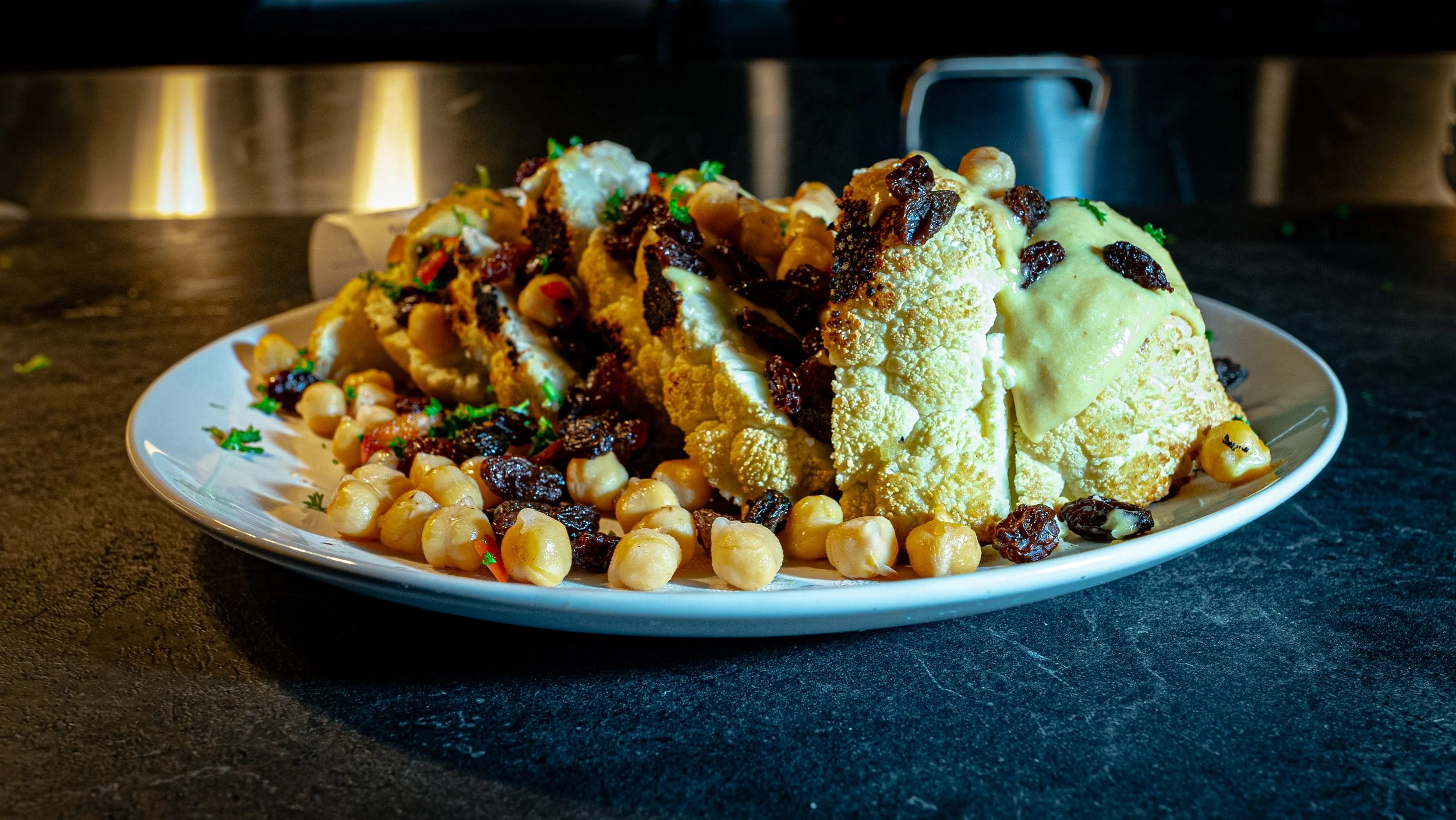 A plate with cooked cauliflower, chickpeas, raisins, herbs, and seasoning on a dark surface.