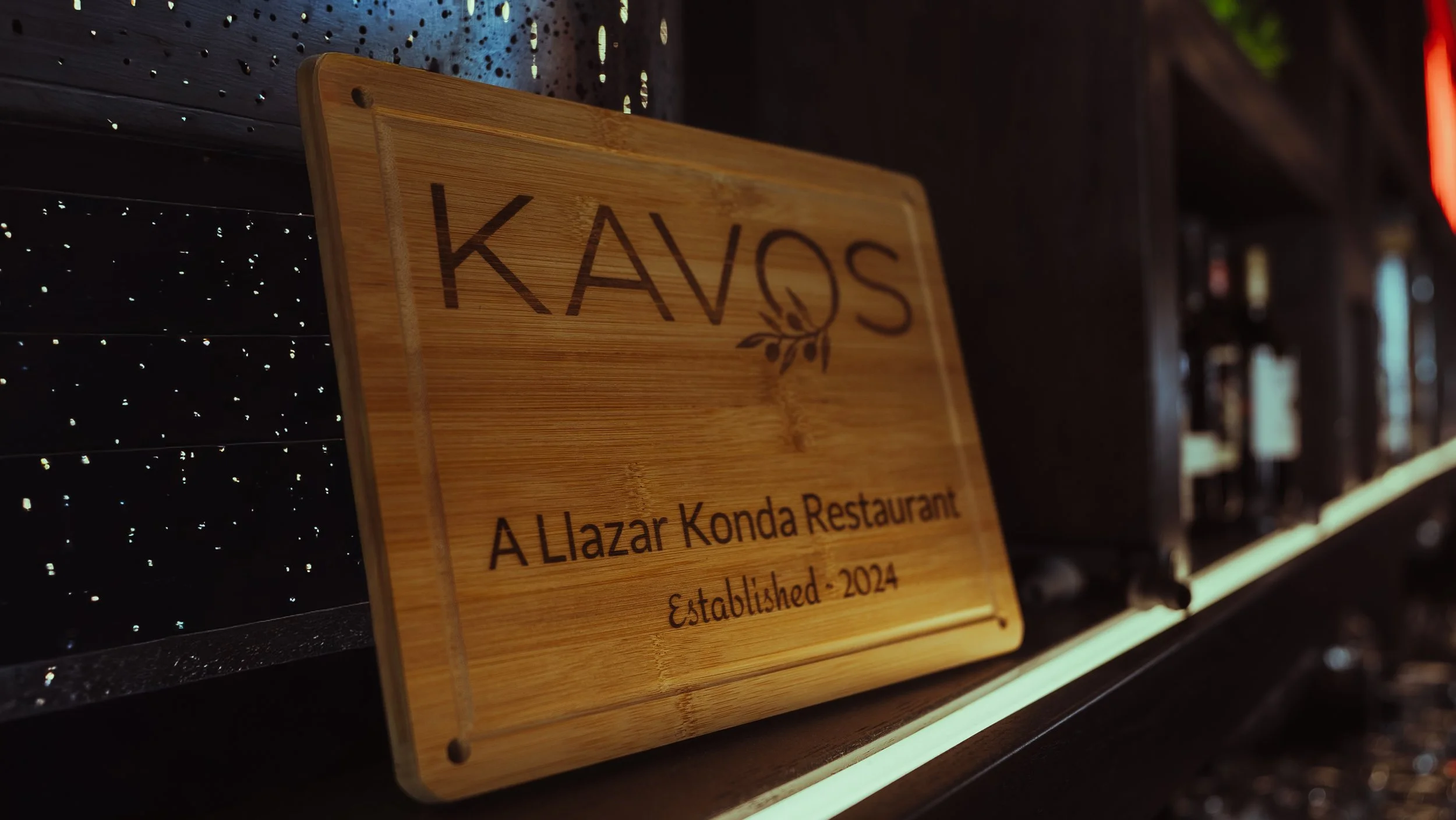 A wooden plaque with the text 'KAYOS' and a tea leaf graphic, indicating it is from A Lizar Konda Restaurant, established in 2024, placed on a dark countertop with a black pegboard background and dim lighting.