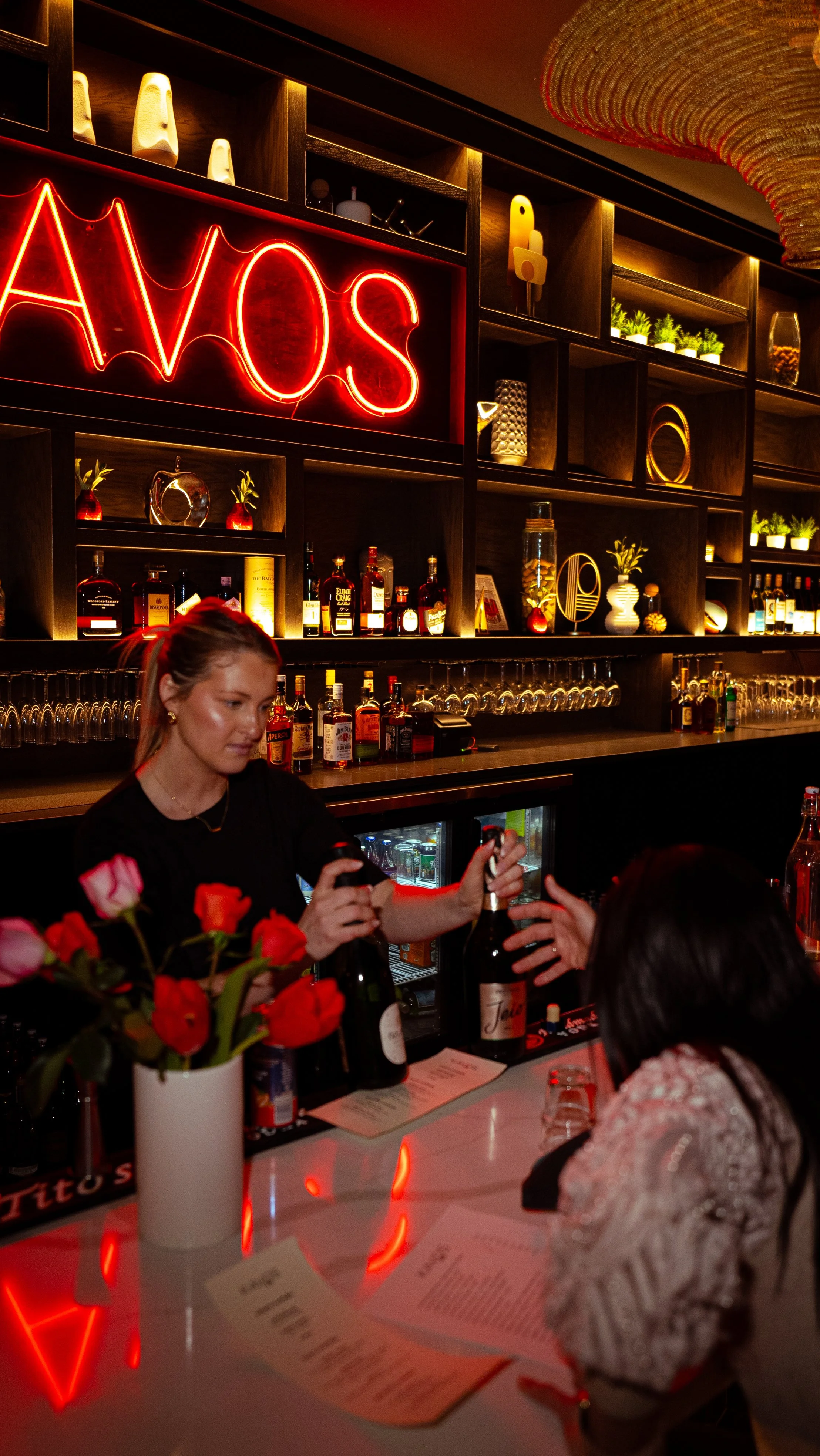 Bar scene with a bartender serving drinks, shelves with bottles and decorative items, and neon sign that says 'WOS' in red.