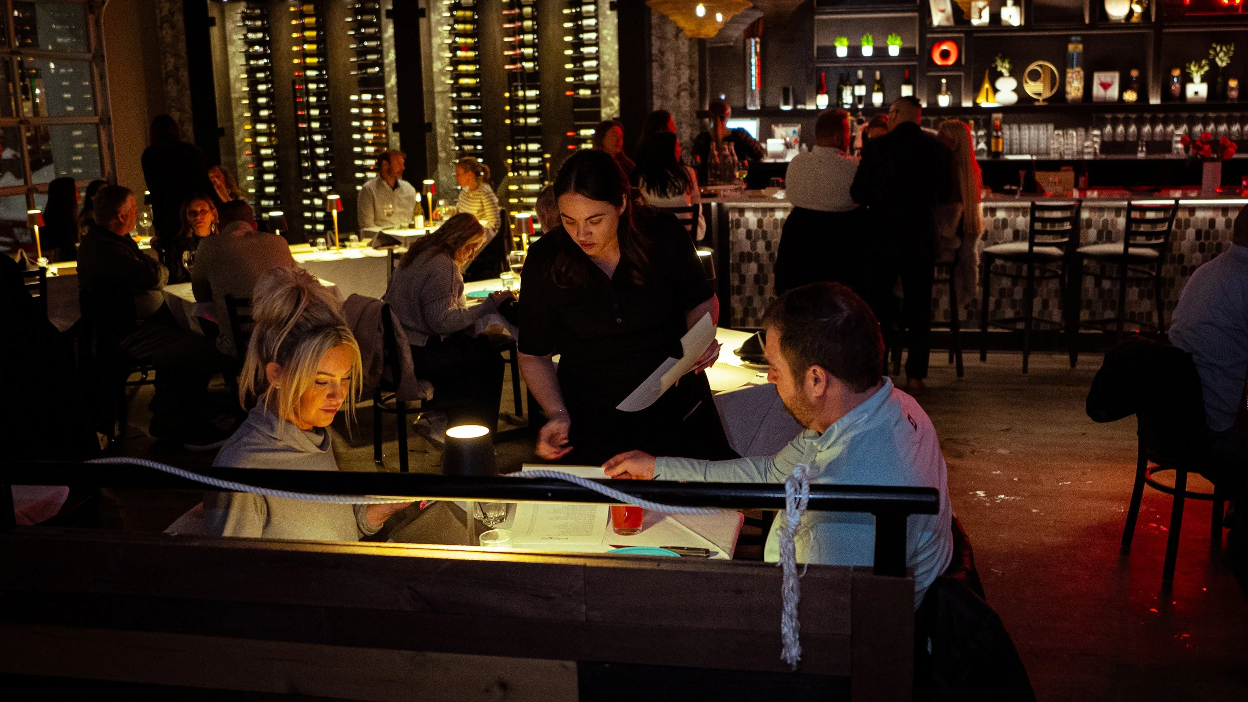 People dining in a dimly lit restaurant with wine bottles on racks and decorative shelves in the background. A waitress serves a seated couple, and other guests are visible at tables.