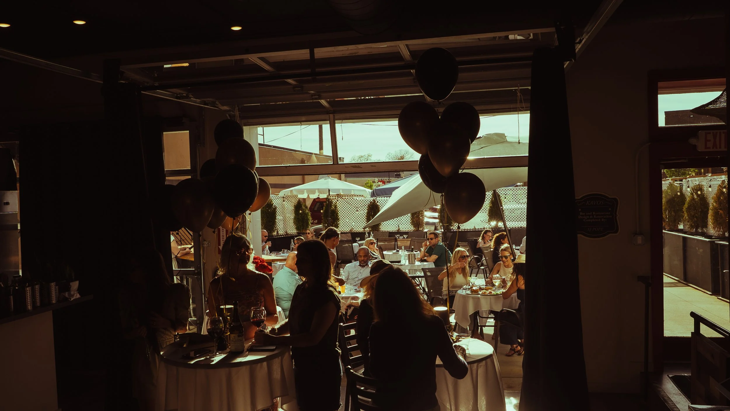 People dining and socializing at an outdoor restaurant patio during sunset, with tables, chairs, and balloons.