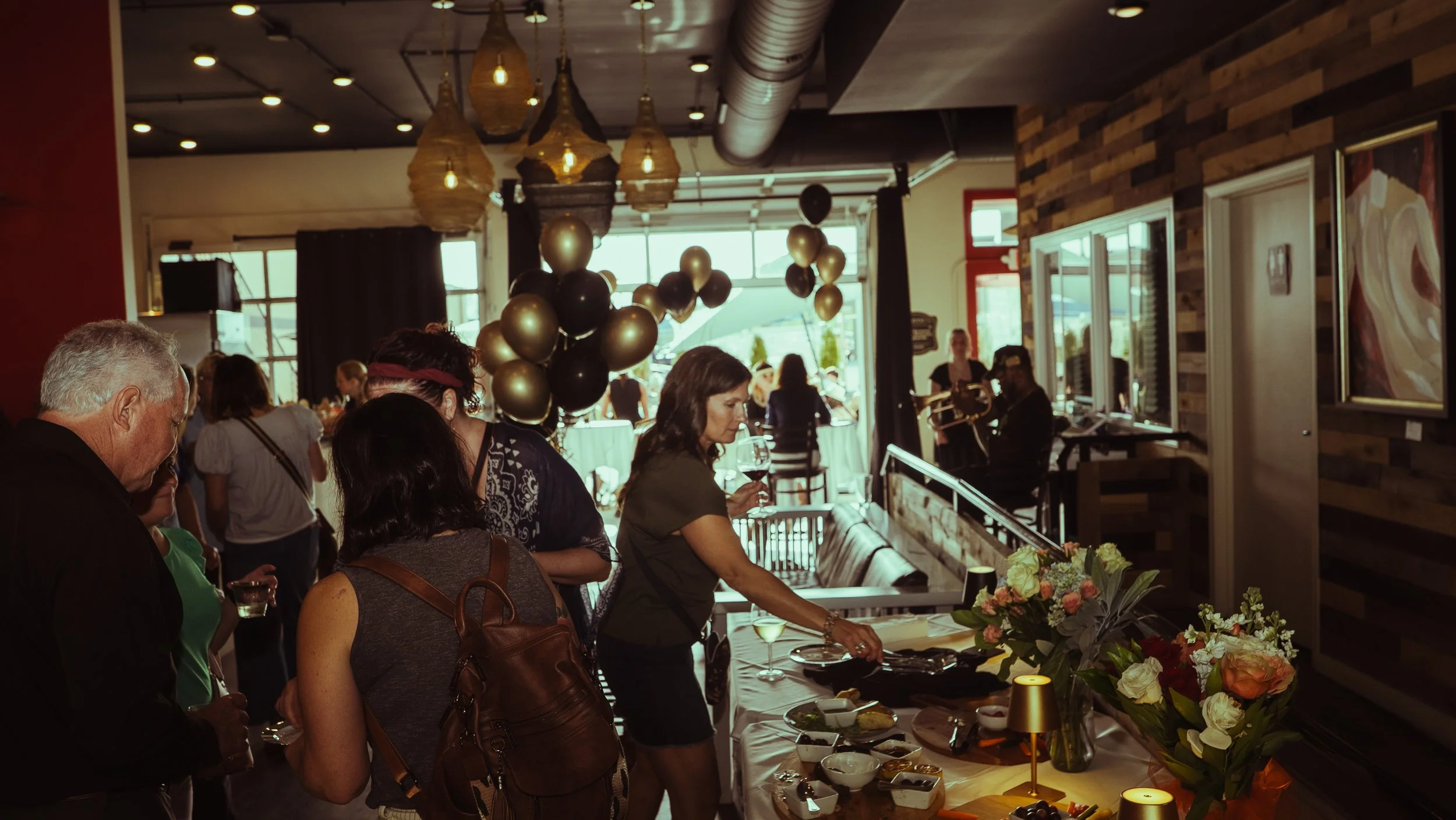 People gathered at a social event inside a restaurant with hanging rope lights and balloons, some of whom are serving or preparing food at a table decorated with flowers.