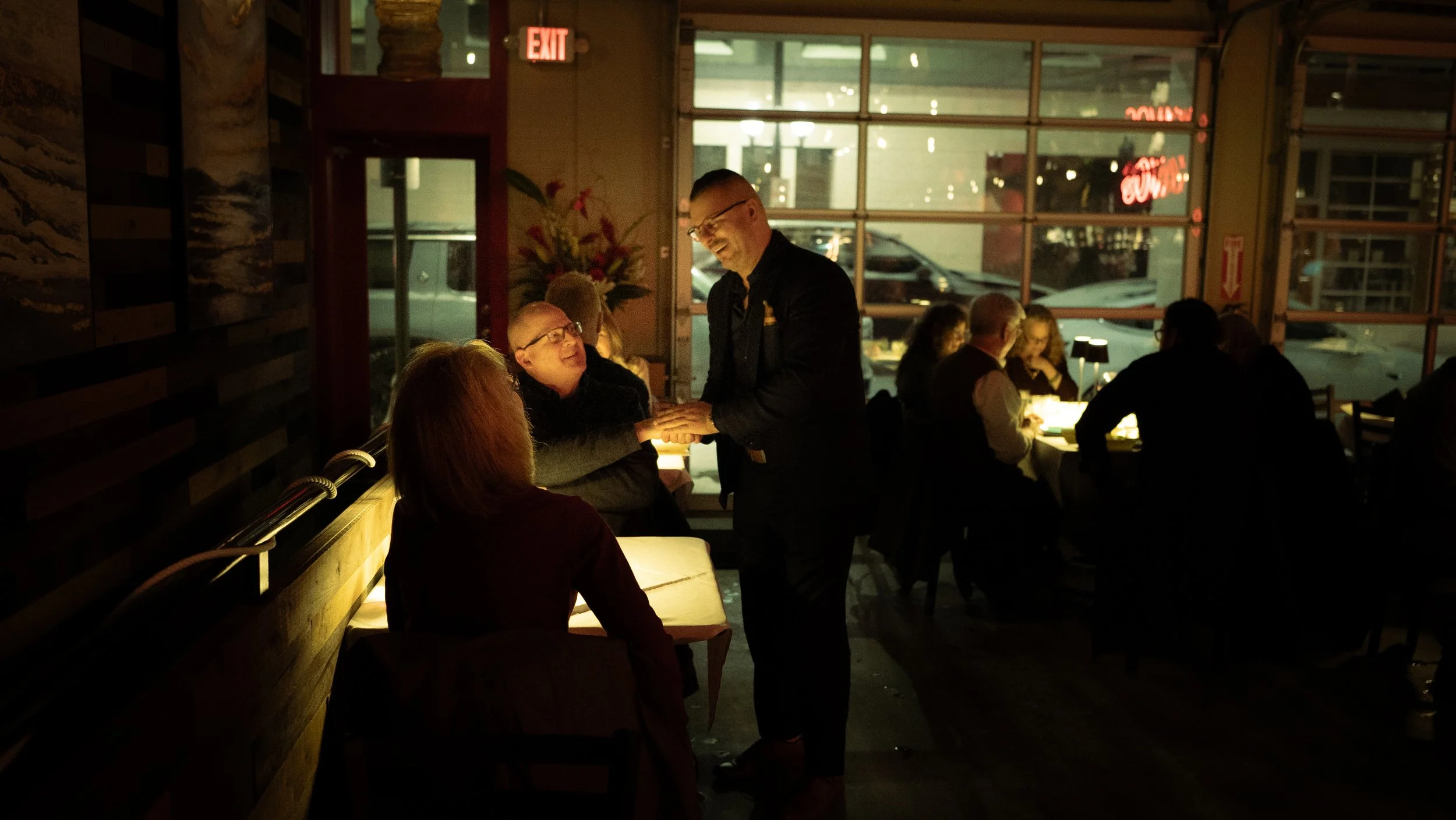 A man in black attire, likely a server, is smiling and interacting with a woman sitting on a bench inside a dimly lit restaurant. Other diners are seated at tables, and large windows behind them reveal the night outside. A neon 'Exit' sign and urban 