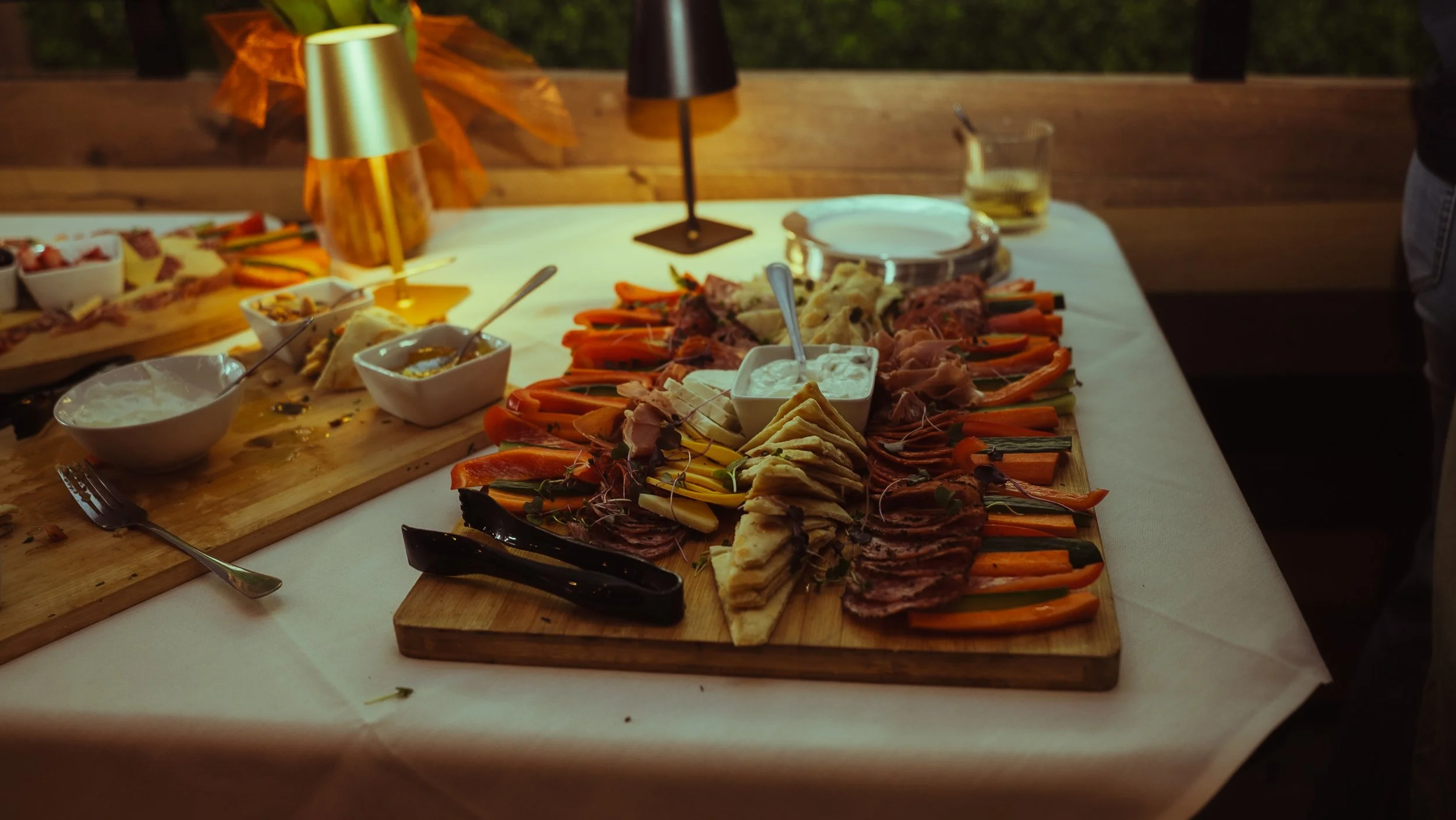 A wooden platter with assorted sliced meats, vegetables, and cheeses, along with various dipping sauces, on a dining table during an outdoor gathering.