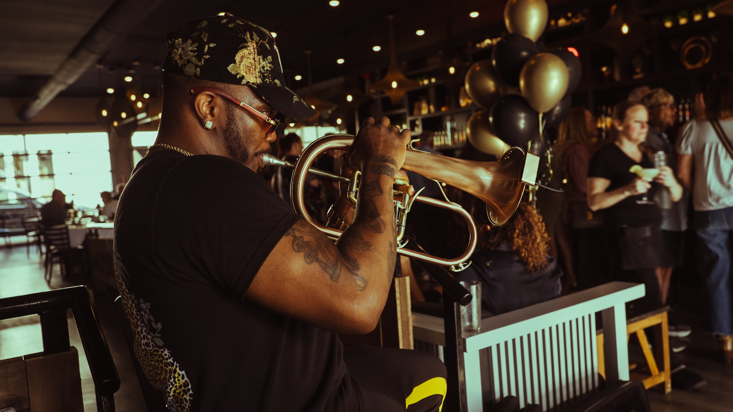 A man wearing sunglasses and a hat playing a trumpet in a lively bar or restaurant with people in the background.