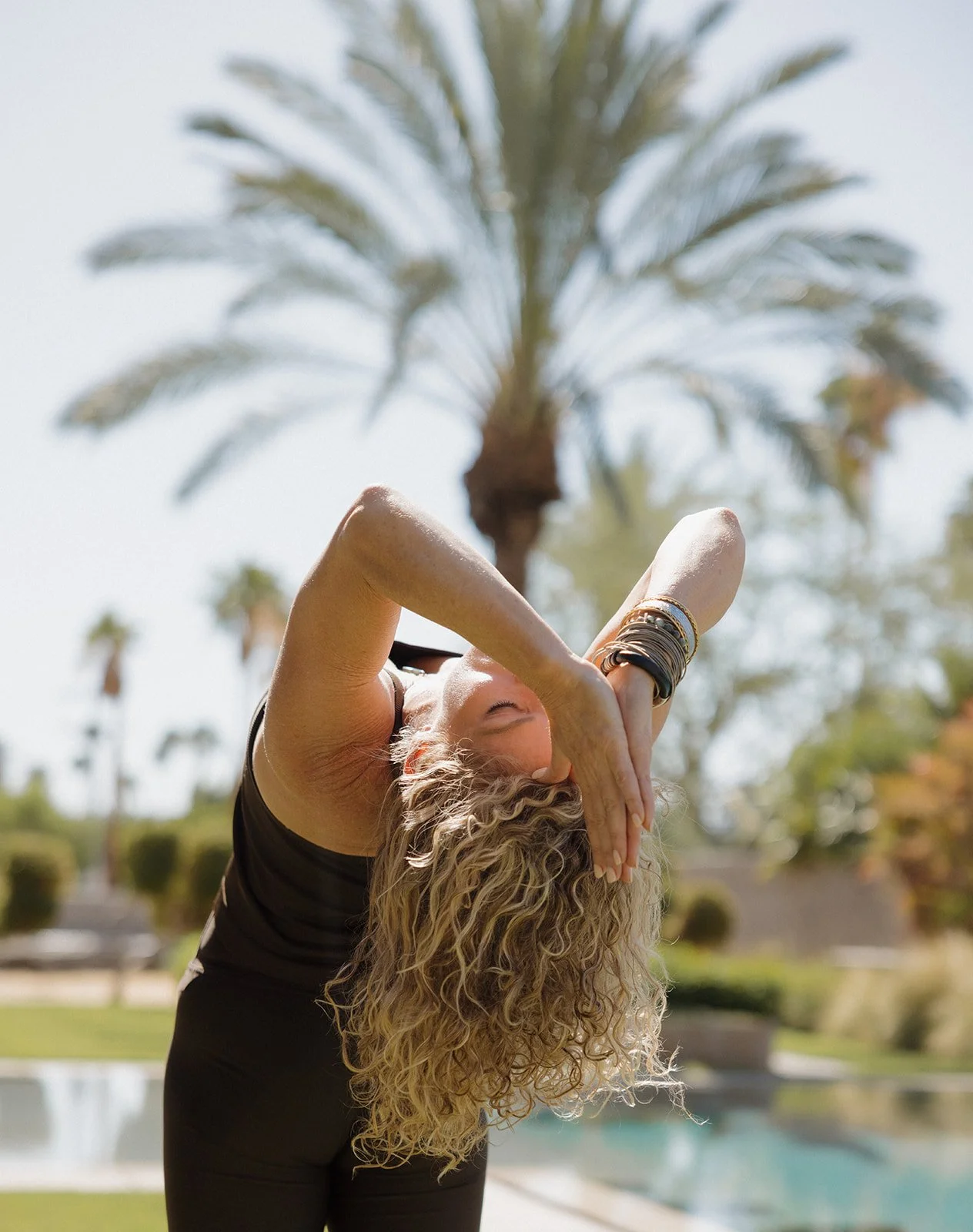 Woman practicing a yoga backbend outdoors near a pool, palm trees overhead.