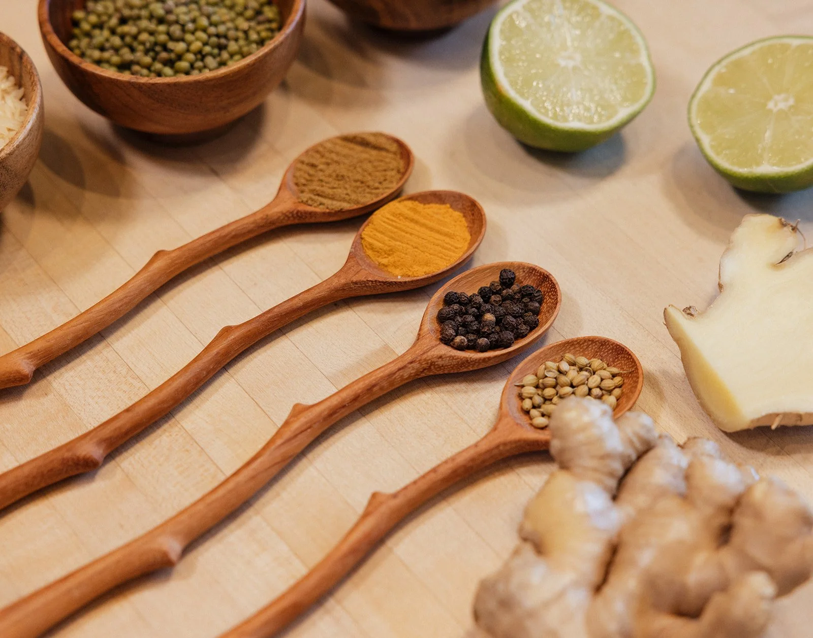 Wooden spoons with ground cumin, turmeric, black peppercorns, and coriander seeds, with lime and ginger.