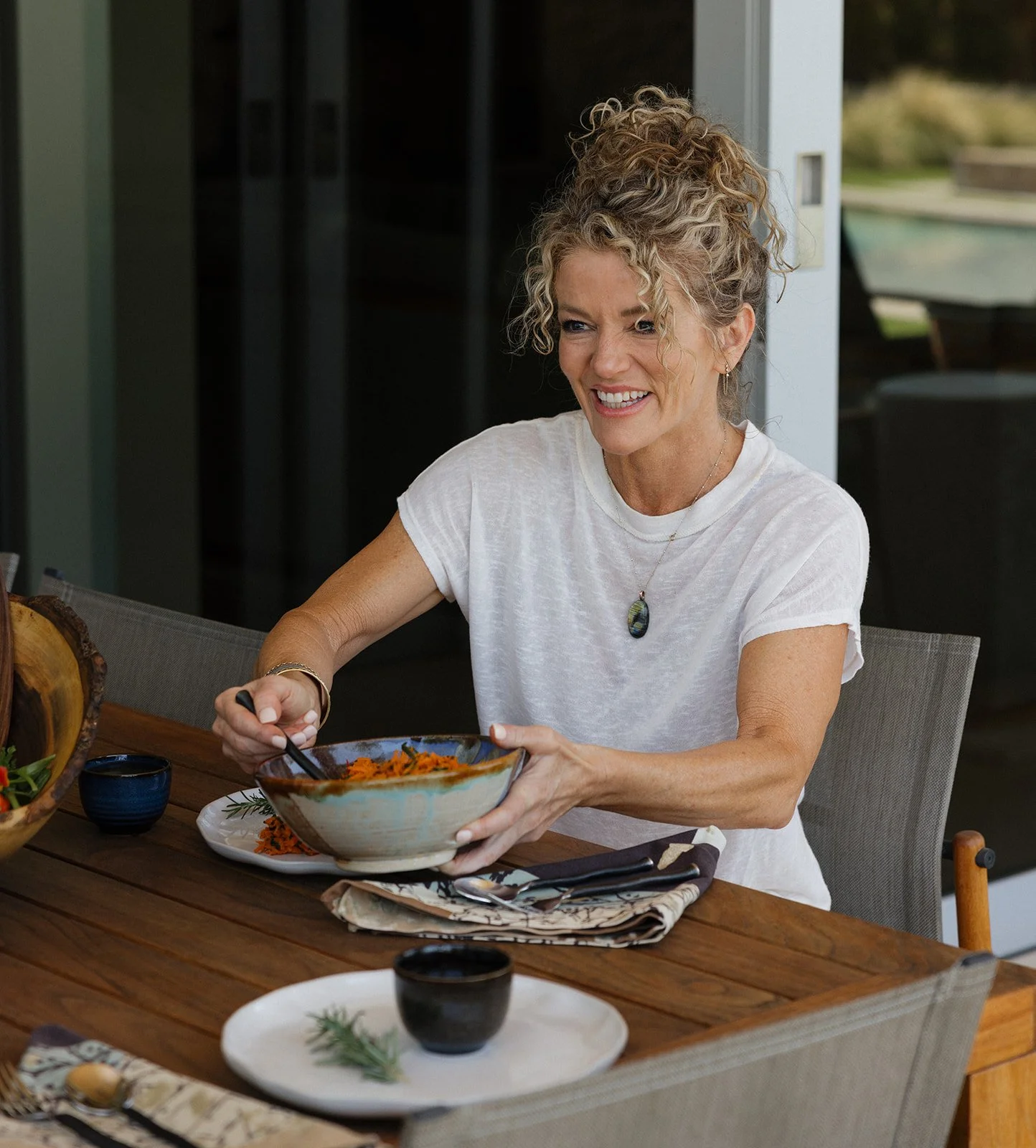 A woman wearing a white t-shirt and necklace, smiling and serving clients at an outdoor dining table at an Ayurveda retreat.