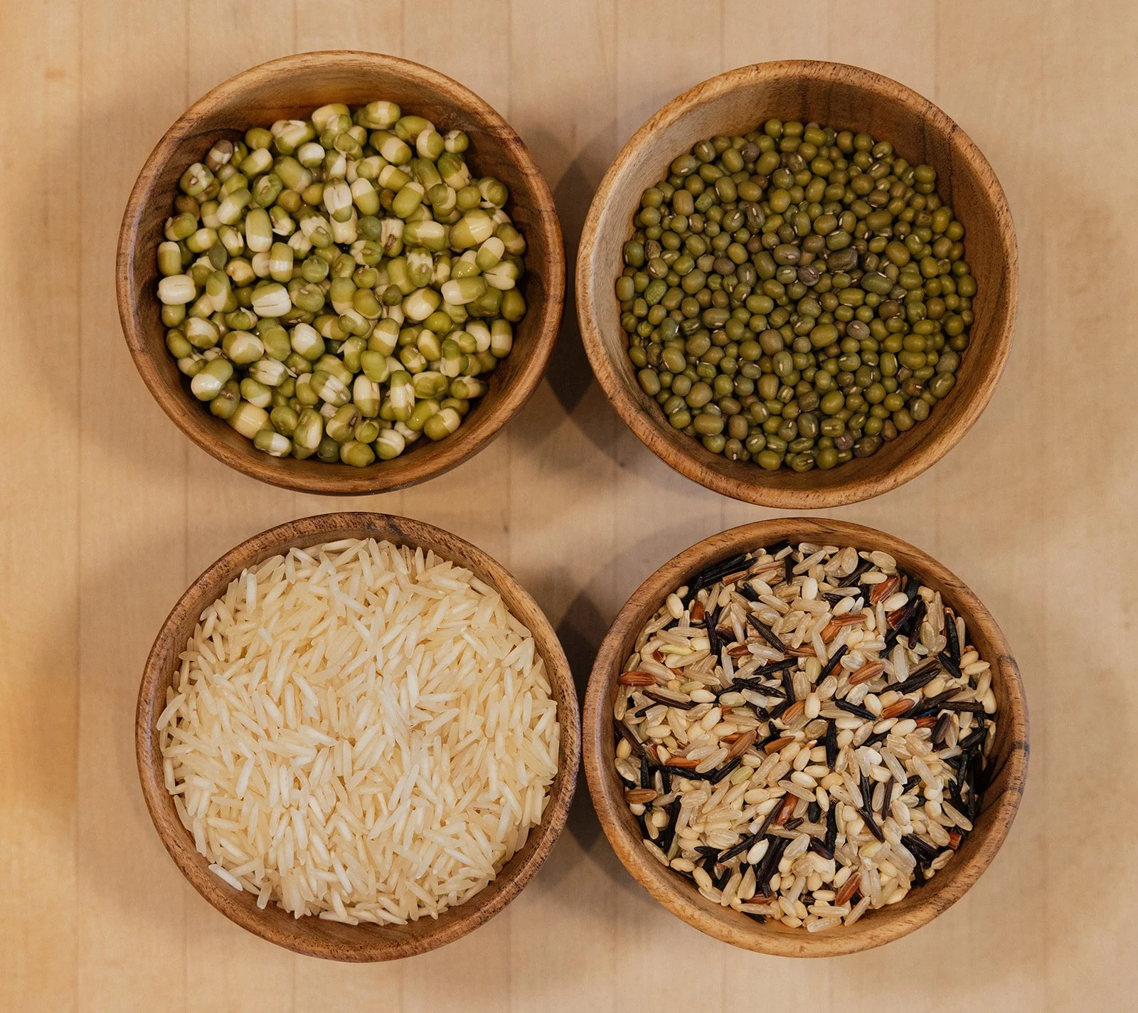 Four round wooden bowls filled with different types of rice and beans for Ayurvedic cooking, arranged on a light wooden surface.