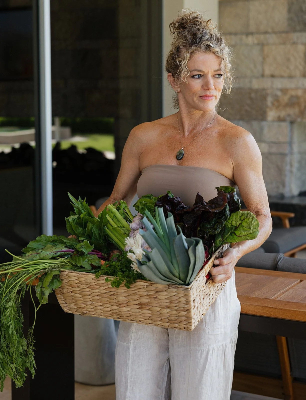 A woman holding a basket of fresh vegetables, including garlic, carrots, and leafy greens, standing outdoors at a Prana and Spice Ayurveda retreat.