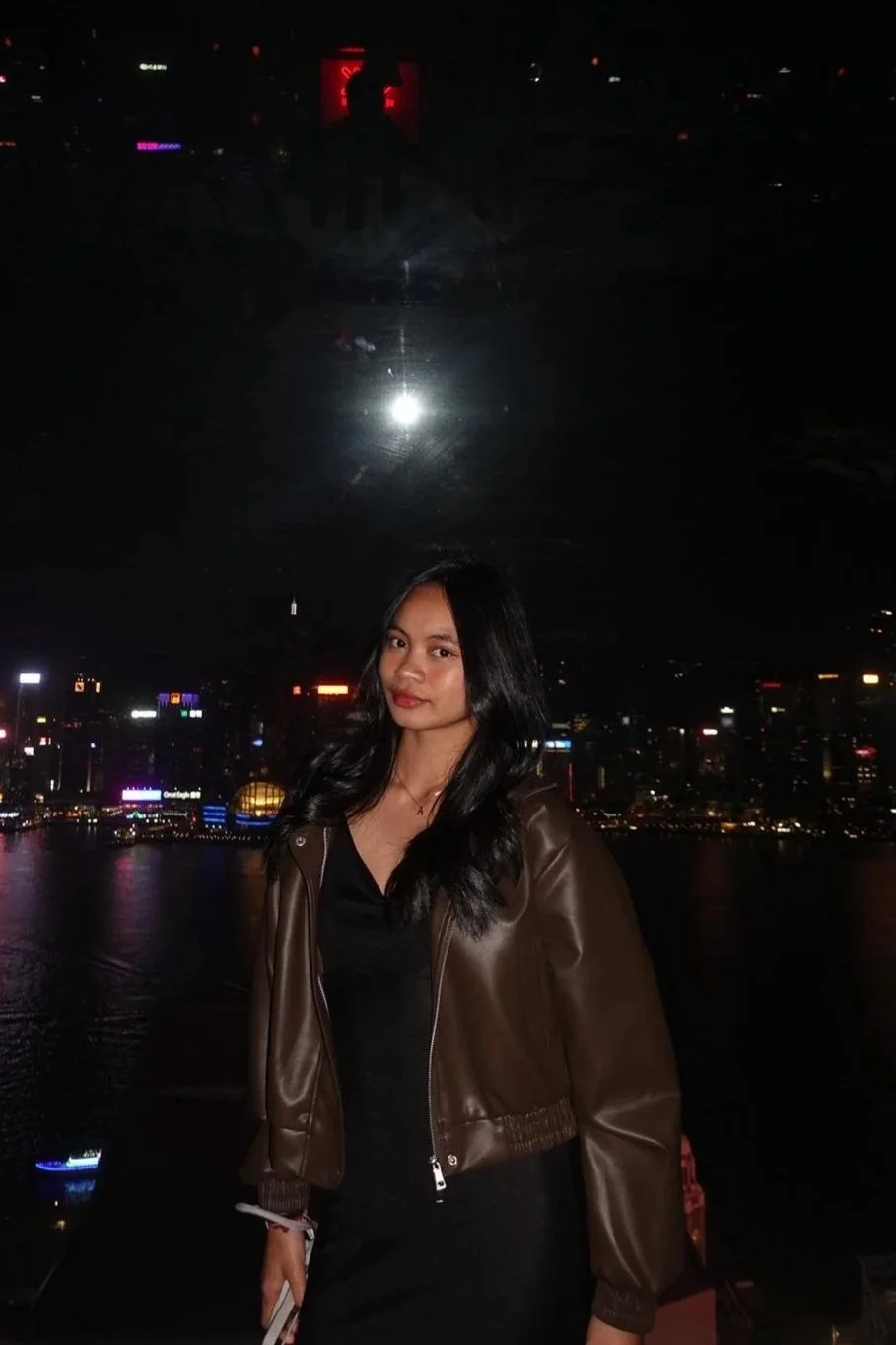 A woman with shoulder-length black hair wearing a brown leather jacket and black top, standing in front of a city skyline at night with illuminated buildings and reflections in the water.