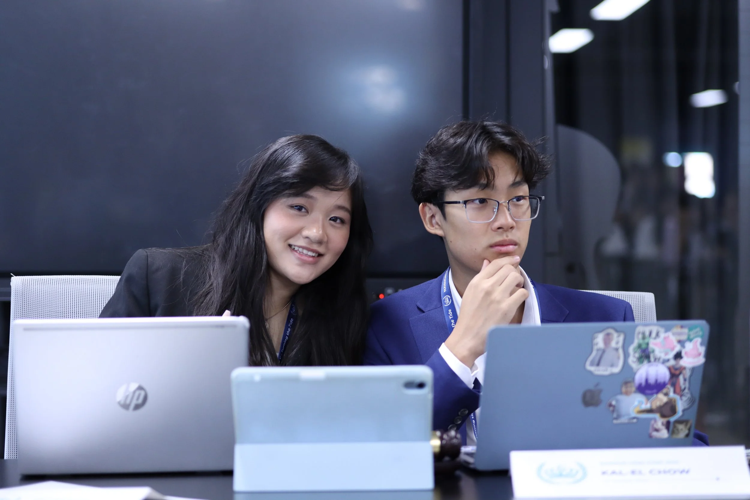 A young woman with long black hair smiling and a young man with glasses and on his chin touching his hand, both sitting at a desk with laptops in an office or conference setting.