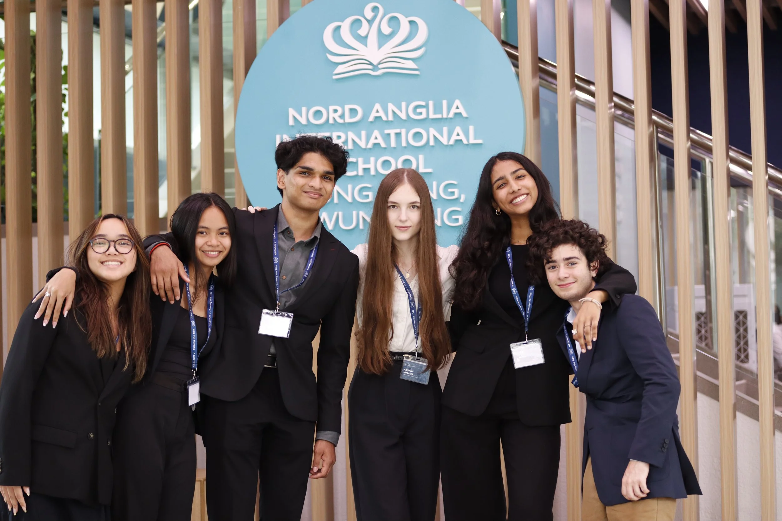 Group of six diverse students smiling and posing together in front of a "Nord Anglia International School" sign at an educational event.