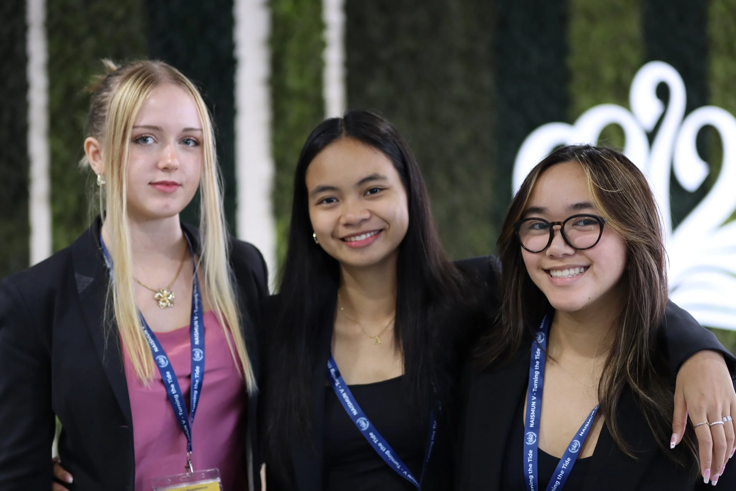 Three young women standing together in a casual indoor setting, smiling at the camera, with a blurred background featuring greenery and a white decorative sign.