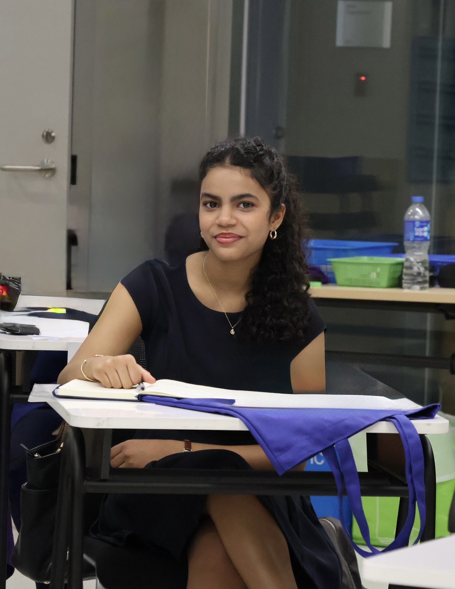 A young woman with curly dark hair, wearing a black dress, sitting at a desk with open notebooks, in an indoor classroom or office setting. There are office supplies on the desk, a water bottle, and some bags hanging on the chair.