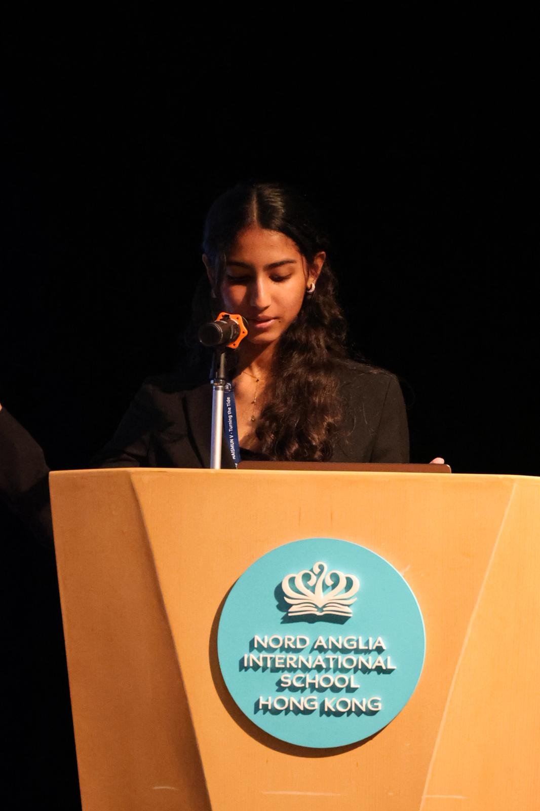A young woman standing behind a podium with Nord Anglia International School Hong Kong logo, speaking or reading into a microphone.