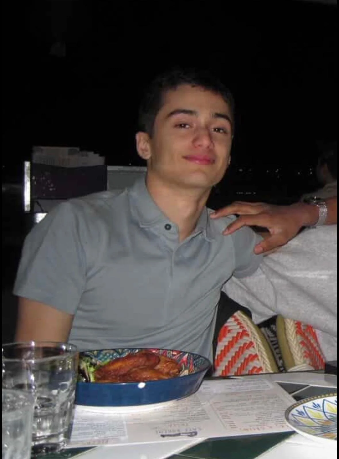 A young man with short dark hair and a light gray shirt, sitting at a dinner table with food and drinks, smiling at the camera in a dark outdoor setting.