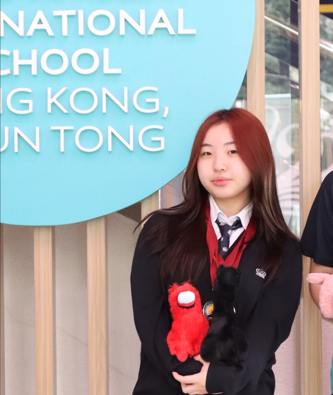 A young woman with long brown hair wearing a school uniform, holding two plush toys, one red and one black, standing in front of a blue sign with white text that reads 'NATIONAL SCHOOL HONG KONG, UN TONG'.