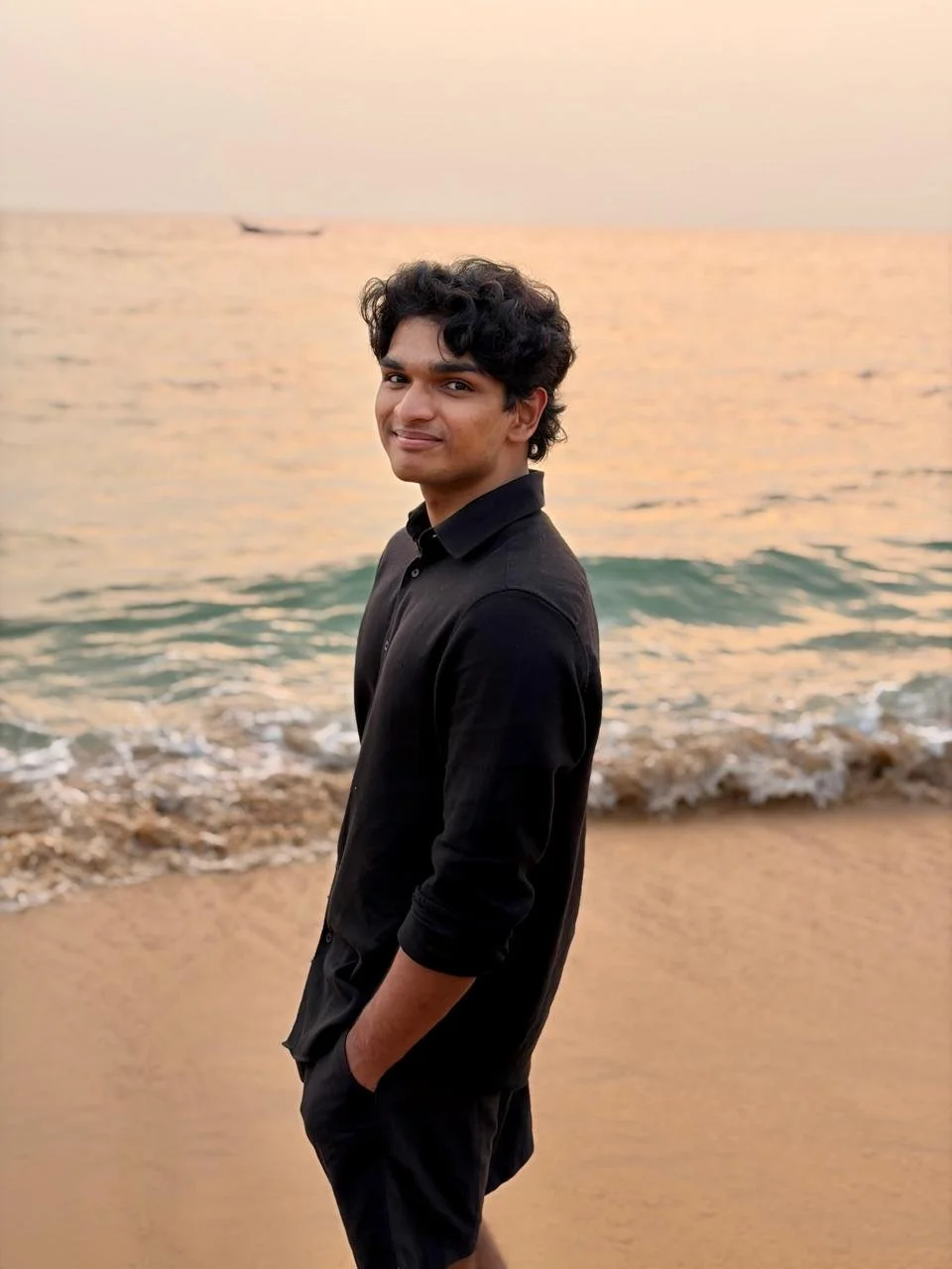 A young man with curly hair standing on the beach during sunset, wearing a black shirt and smiling.