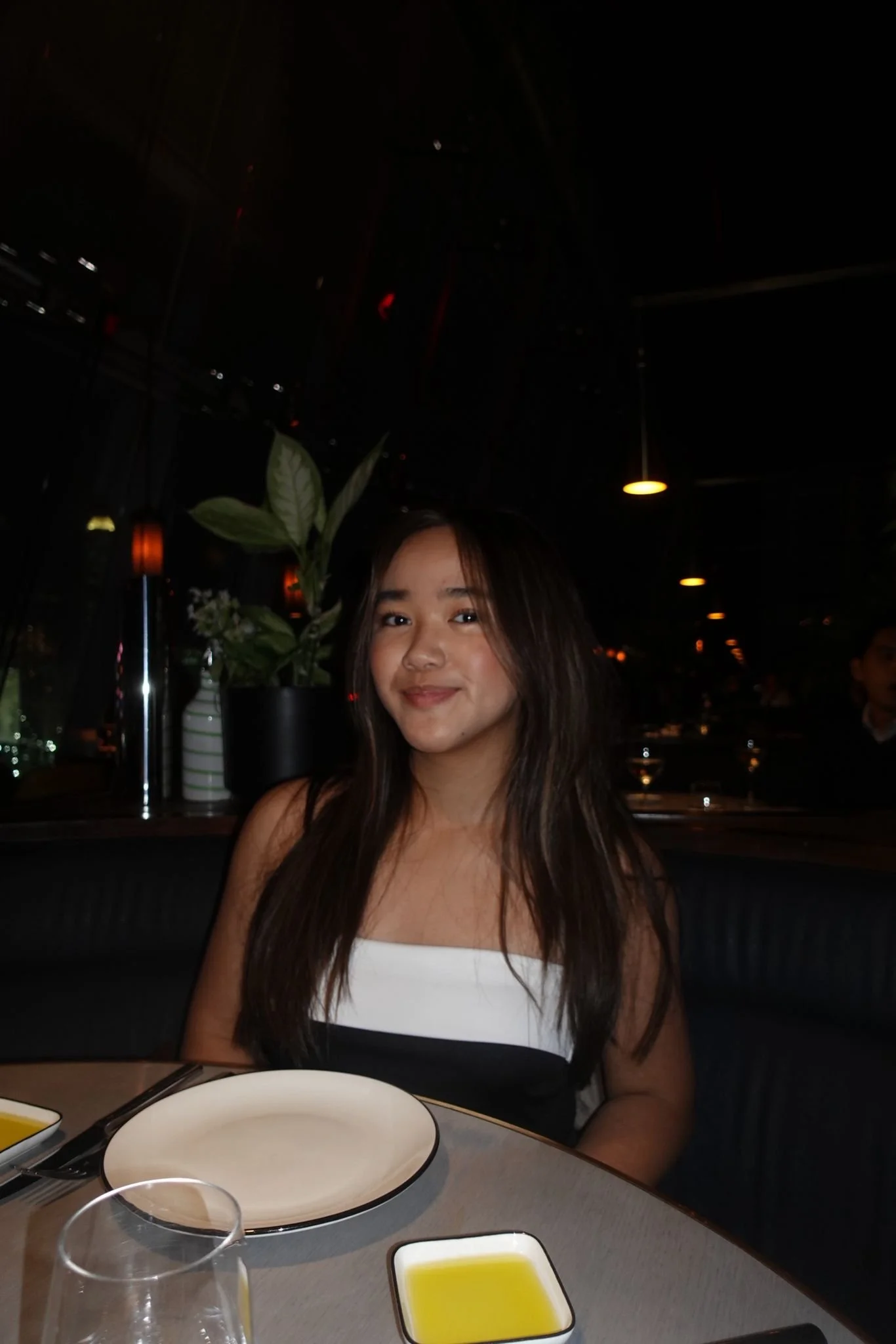 A young woman with long dark hair sitting at a table in a dimly lit restaurant, smiling at the camera. On the table are empty plates, a glass of water, and a small dish of olive oil.