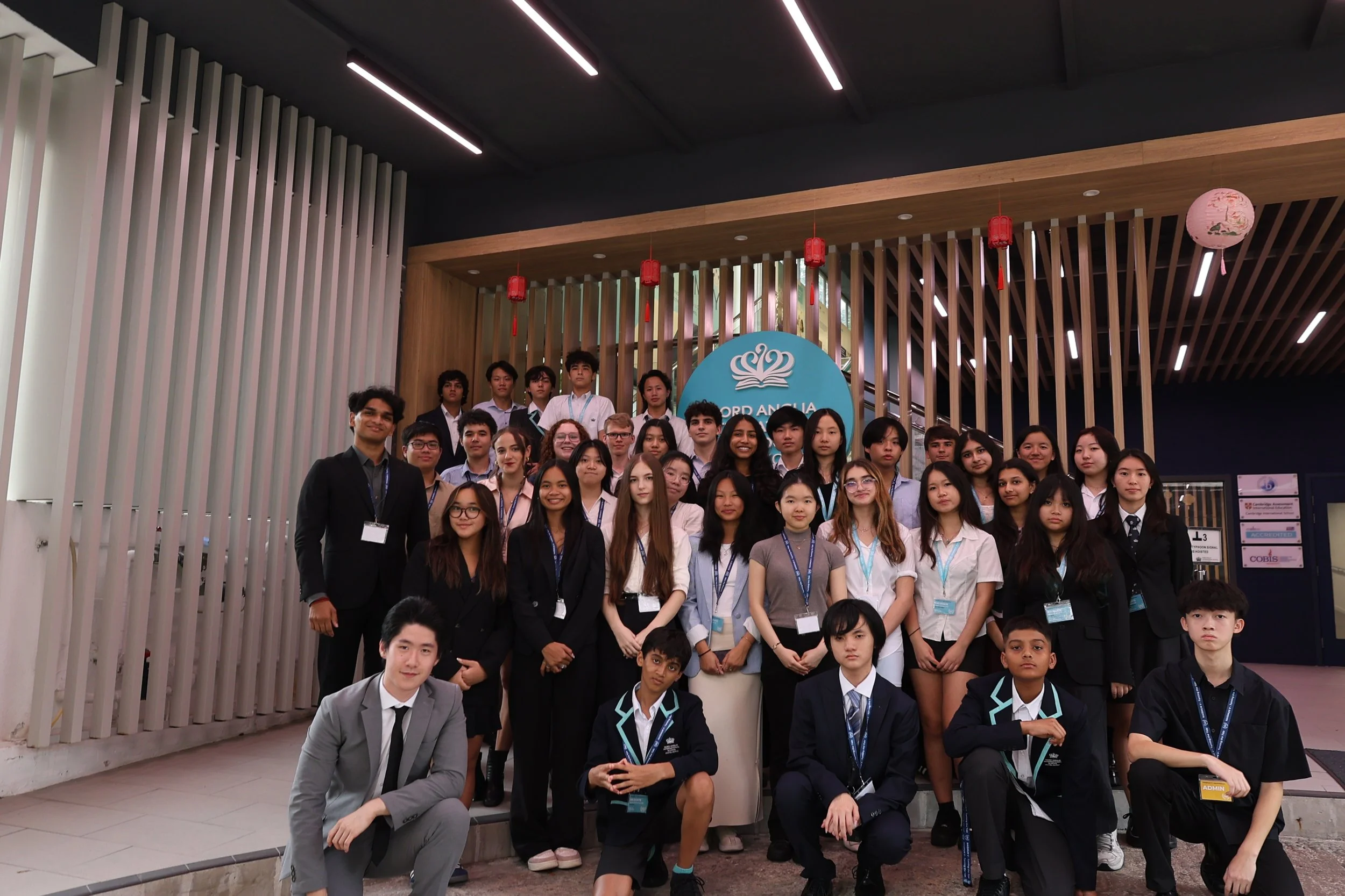 Group of diverse young students and teachers gathered for a photo in front of a sign that reads 'Oxford Anglia' at an indoor event, with the background featuring modern wall and ceiling design with hanging lanterns.