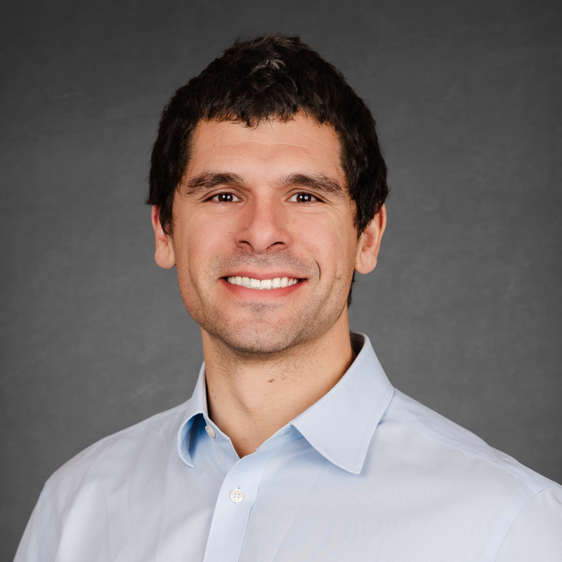 A smiling man with dark hair in a light blue collared shirt against a gray background.