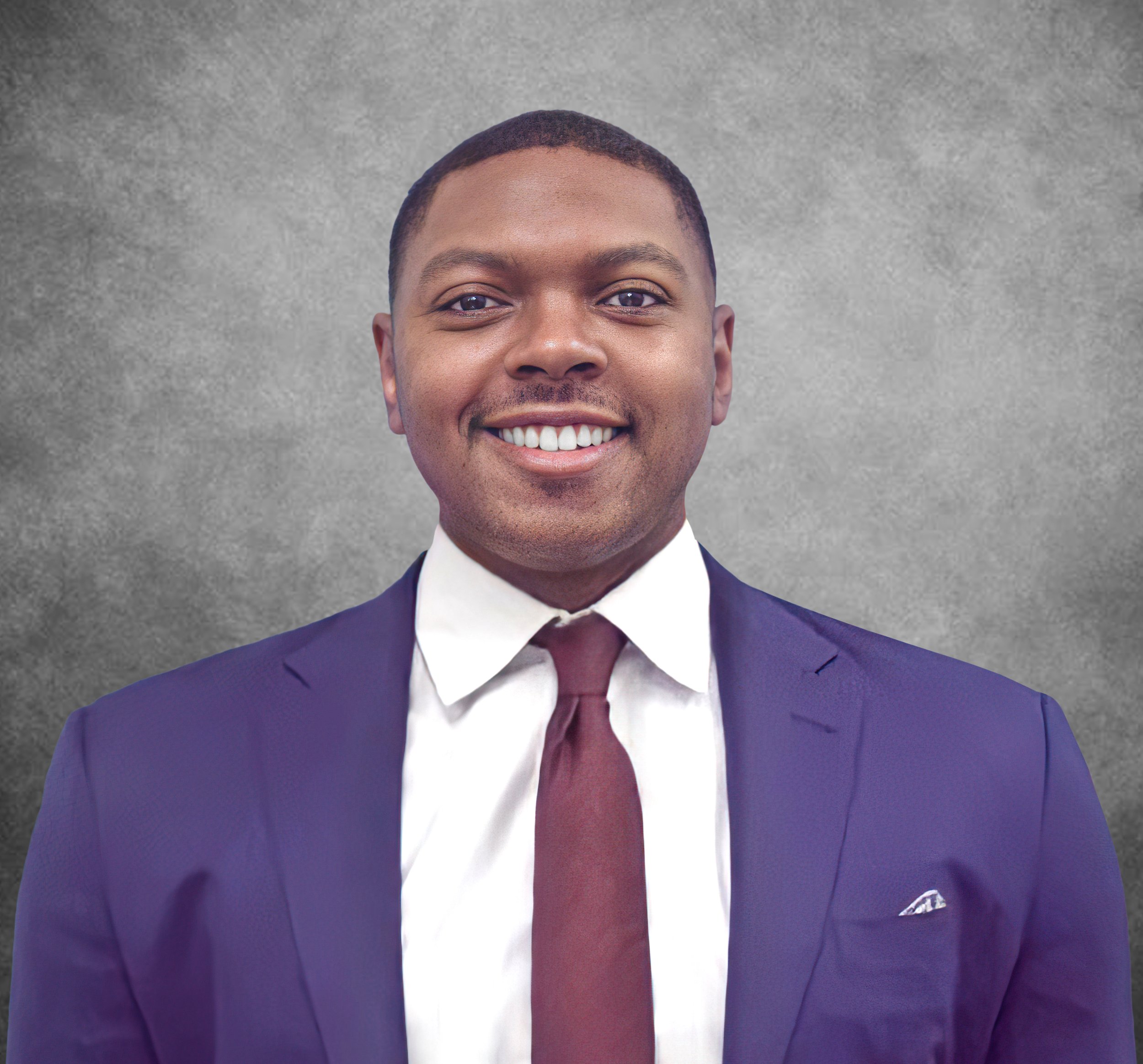 Close-up of a smiling man in a dark blue suit, white shirt, and maroon tie, standing against a gray background.