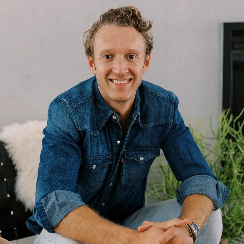 A smiling man with curly hair and a denim shirt, sitting on a couch in a modern living room with green plants.