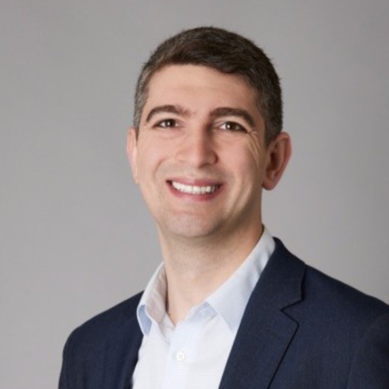 Professional headshot of a smiling man with short dark hair, wearing a navy suit and white shirt against a gray background.
