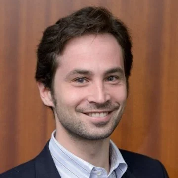A smiling man with dark hair and facial hair, wearing a suit jacket over a collared shirt, standing in front of a wood-paneled background.