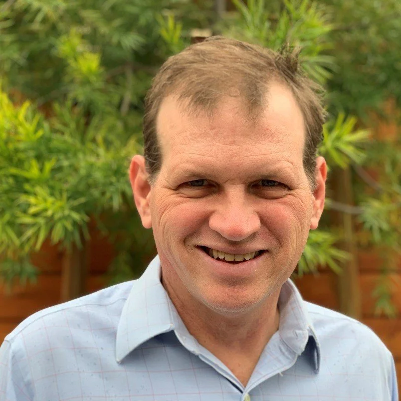 Smiling man with short light brown hair, wearing a light blue collared shirt, outdoors with green foliage and a fence in the background.