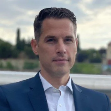 A man in a suit standing outdoors with a cloudy sky and greenery in the background.