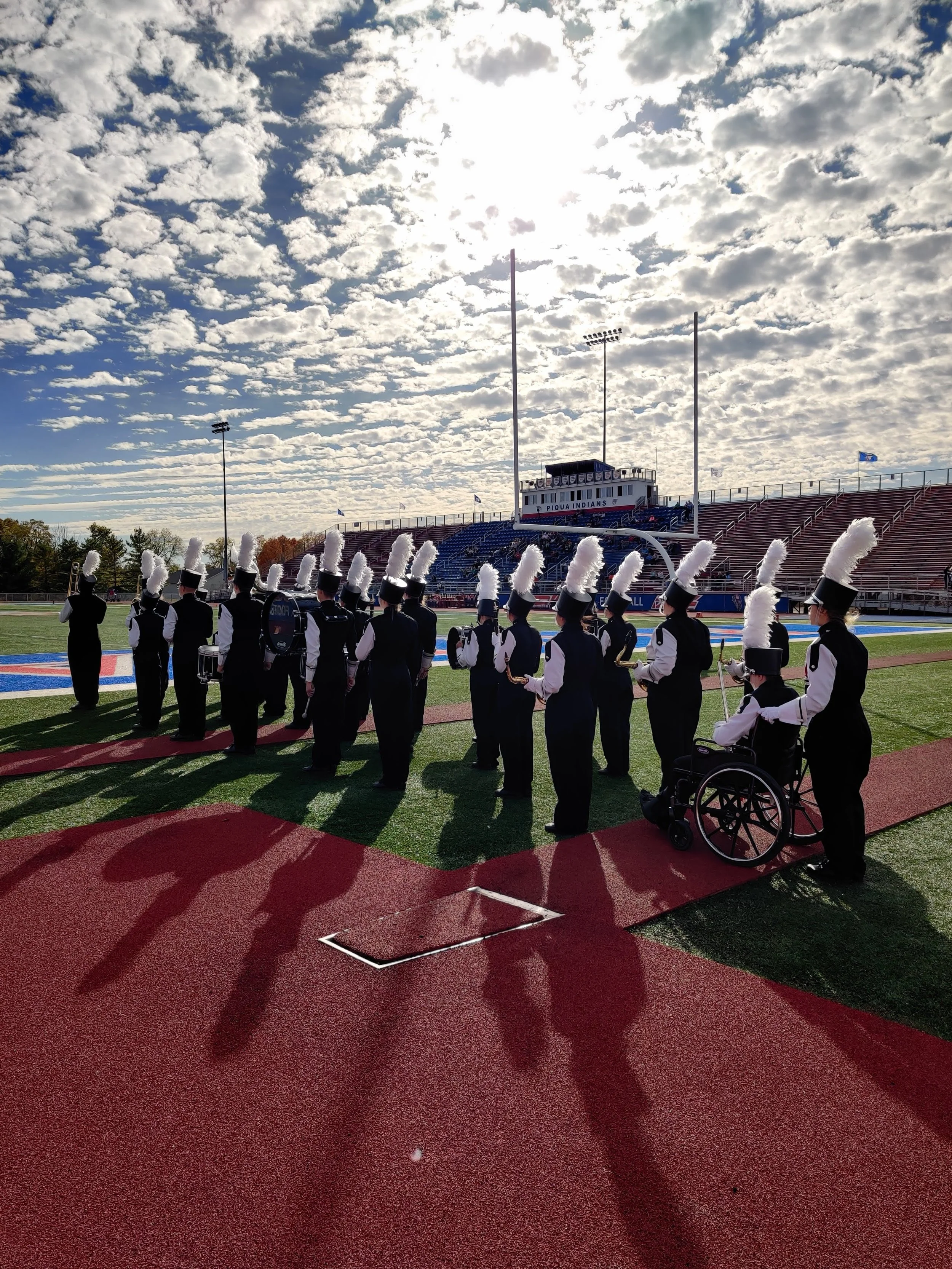 A marching band in uniform with tall hats and feather plumes, standing on a football field. In the background, there is a stadium with a sign that reads "PIQUA INDIANS" and empty bleachers. The sky has scattered clouds with the sun shining brightly. There are several flags visible near the stadium.