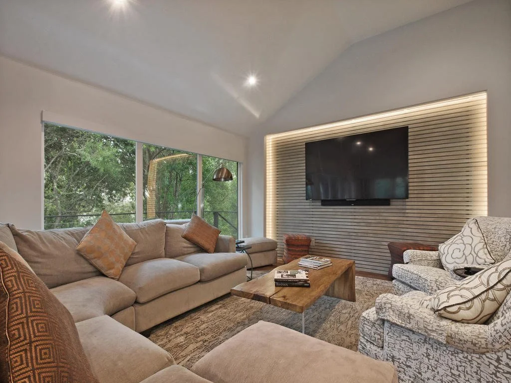 Living room with large beige sofa, armchairs with patterned fabric, wooden coffee table, mounted flat-screen TV on a wood-paneled accent wall, large windows with trees outside, and modern lighting.