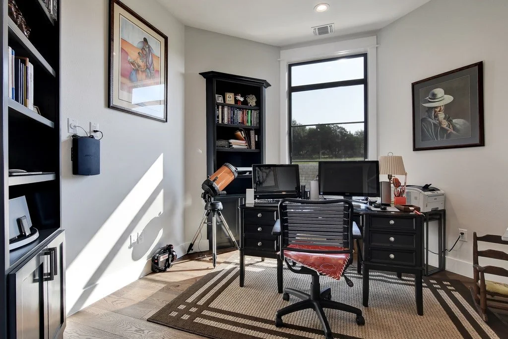 Home office with two computer monitors, a desk with office supplies, a black chair, a telescope, and framed artwork on the walls.