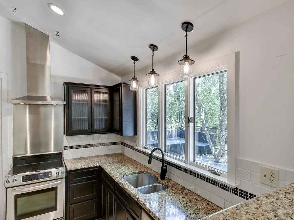 Kitchen with black cabinets, granite countertops, stainless steel stove, large window with a view of a deck area with trees outside, and three hanging pendant lights.