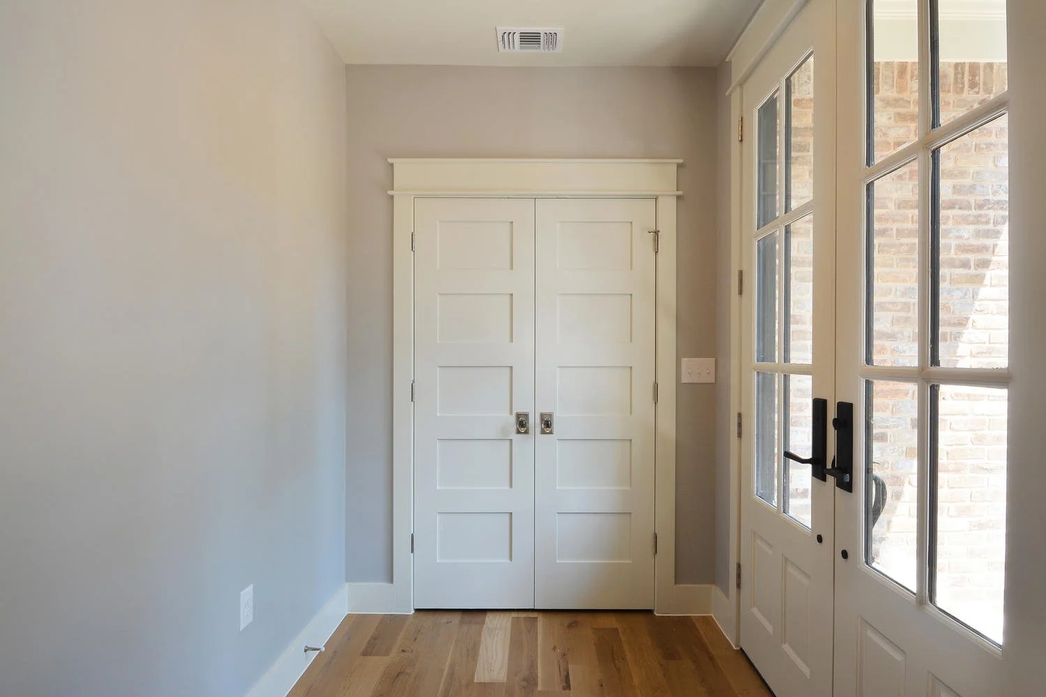 A small entryway or mudroom with a white double closet door, beige walls, a hardwood floor, and a white door with glass panes leading outside.