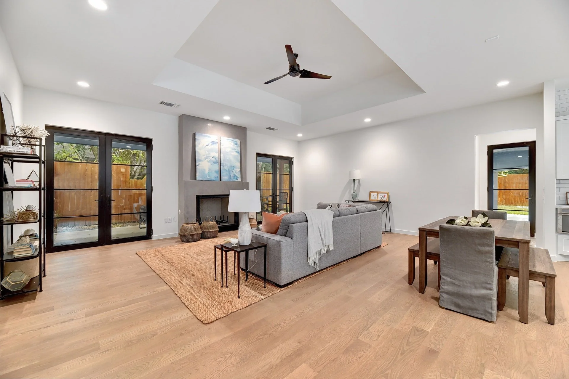 Spacious living room with hardwood floors, a gray sofa, a wooden dining table with chairs, a black metal bookshelf, and a stone fireplace with artwork above it. Large glass doors lead outside, and there is a ceiling fan and recessed lighting.