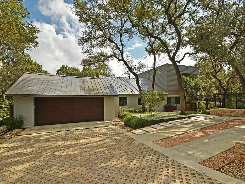 Modern house with a metal roof, attached garage, a landscaped front yard with trees, small shrubs, a stone wall, and a decorative concrete driveway.