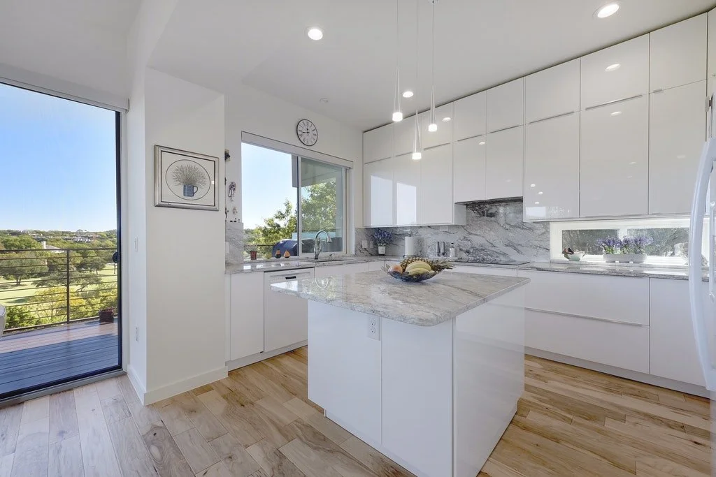 Modern white kitchen with marble countertops, a small island, and wooden flooring, with a balcony door showing outdoor trees and a clear sky.