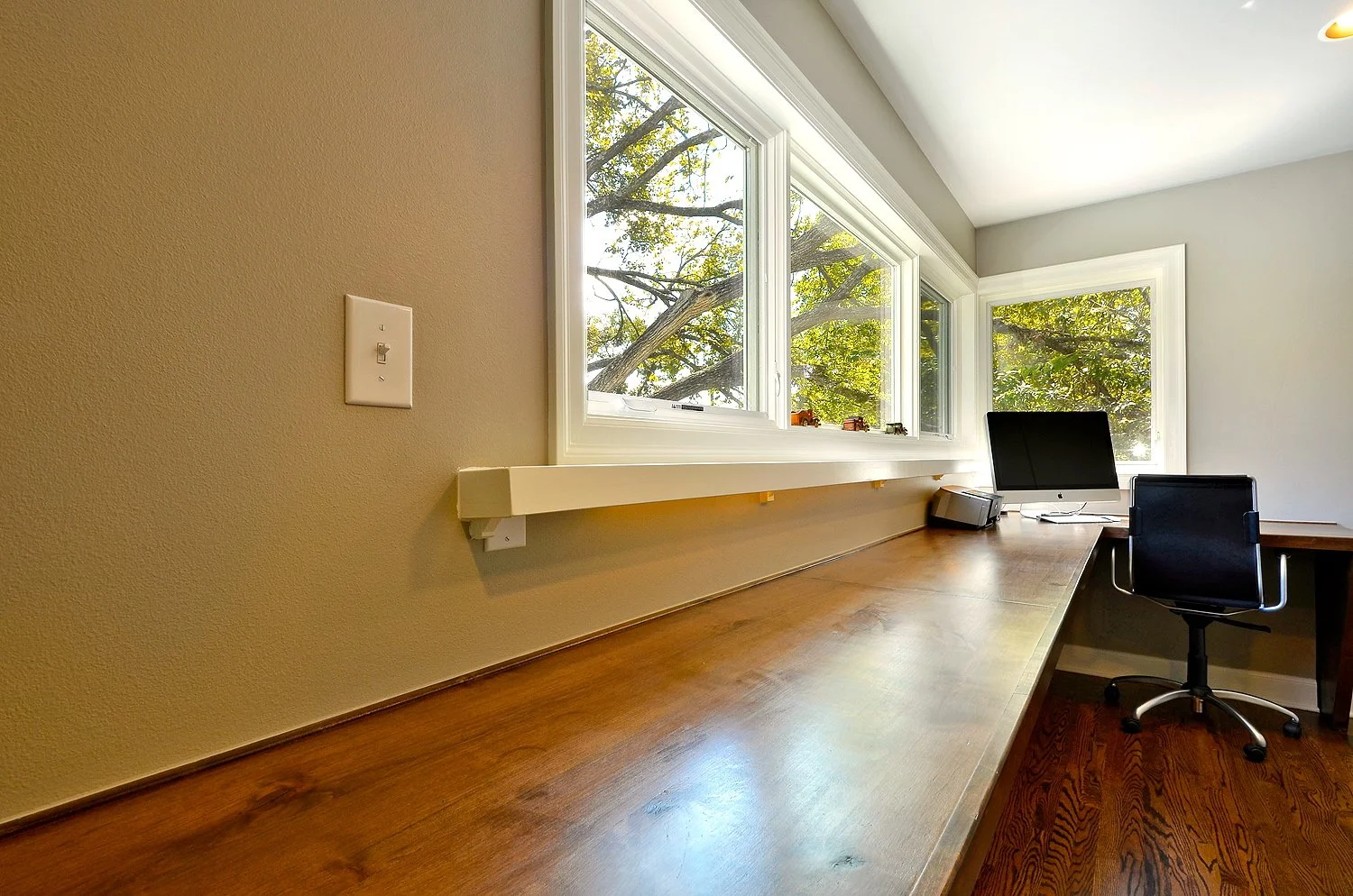 Home office with large windows showcasing trees outside, wooden desk, black office chair, computer monitor, and small electronic device.