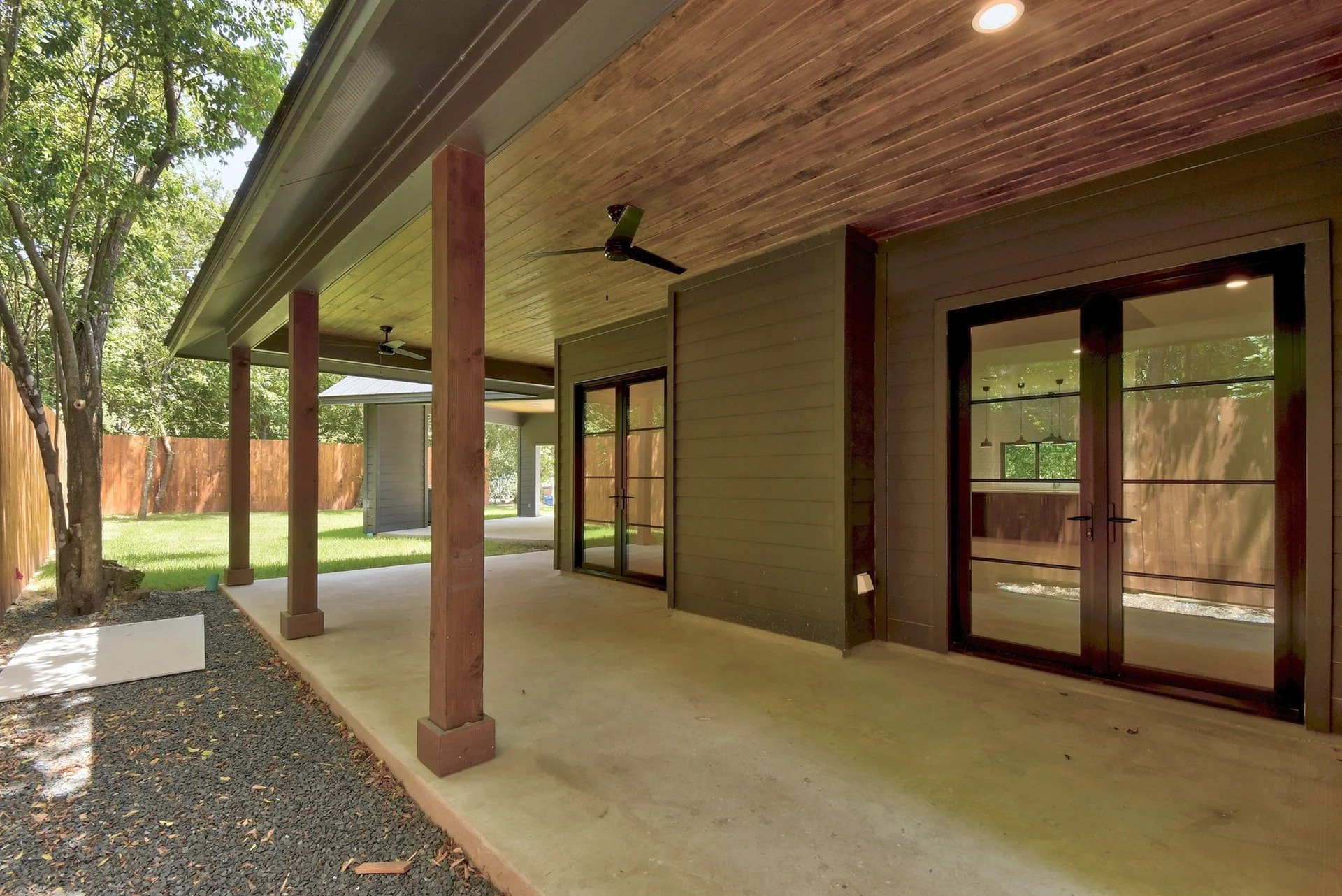 Covered patio area with wooden ceiling, ceiling fans, and large glass sliding doors leading inside, surrounded by green trees and a wooden fence.