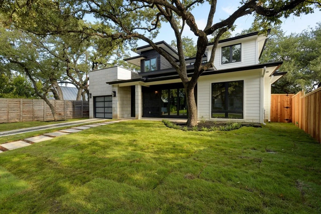 Modern two-story house with white exterior, black accents, large windows, a front yard with a tree, a concrete pathway, and a wooden privacy fence.