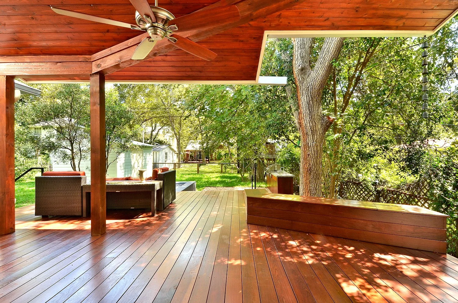 A wooden porch with outdoor furniture, ceiling fan, and a view of trees and a backyard.