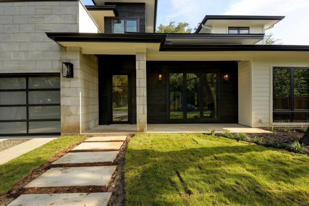 Front exterior of a modern house with a stone and wooden facade, large glass sliding doors, and a well-maintained lawn with a stepping stone pathway leading to the entrance.
