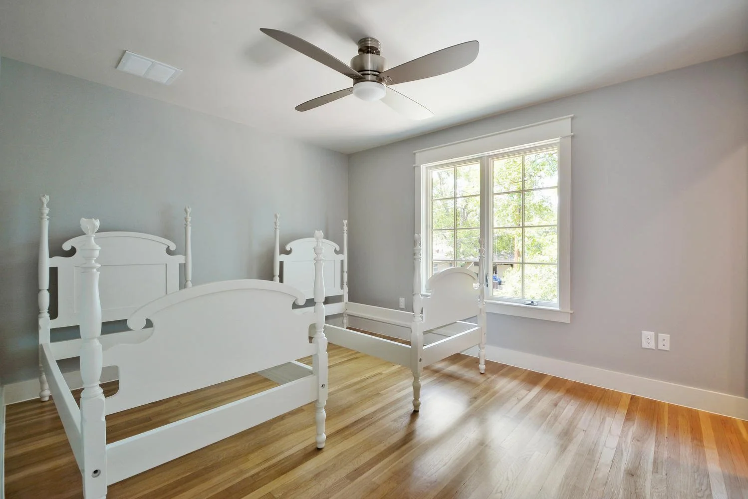 Empty bedroom with two white bed frames, large double window, ceiling fan, hardwood floor, and light gray walls.
