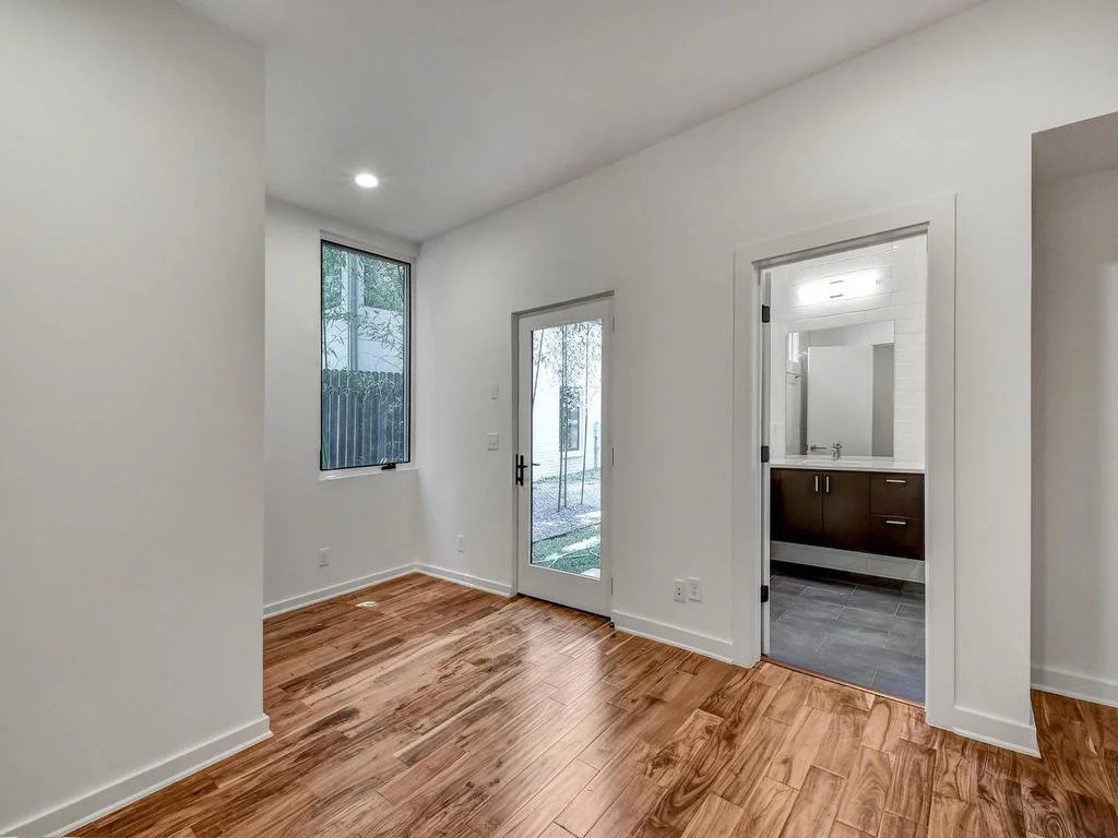 Empty room with hardwood floors, a window, a glass door leading outside, and a bathroom with dark floors and vanity visible through the doorway.