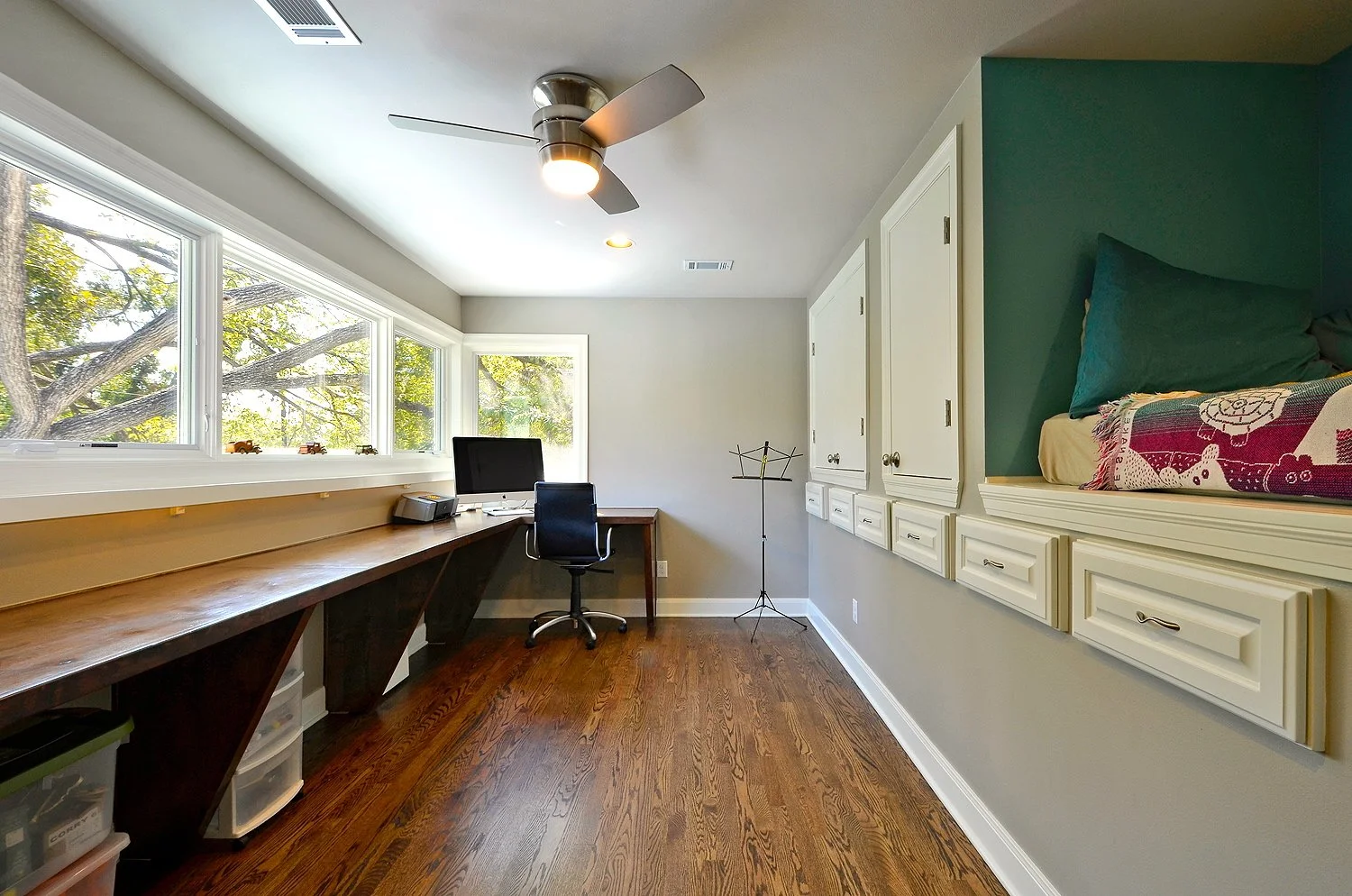 A bright home office with a large window, wooden desk, office chair, computer, and a built-in window seat with cushions.